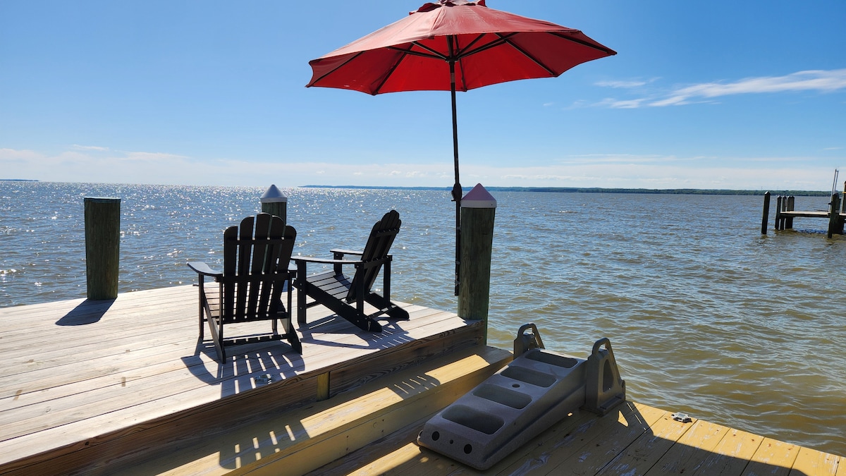 A private pier features two wooden lounge chairs under a red umbrella, overlooking the Potomac River. Sunlight glimmers on the water, and a thoughtfully placed platform is visible nearby, providing additional space for relaxation.
