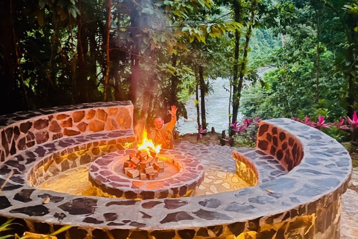 A circular stone fire pit is surrounded by natural stone seating, nestled among lush greenery. Smoke rises from the center, blending with the serene background of trees and visible river, creating a cozy and inviting outdoor space.
