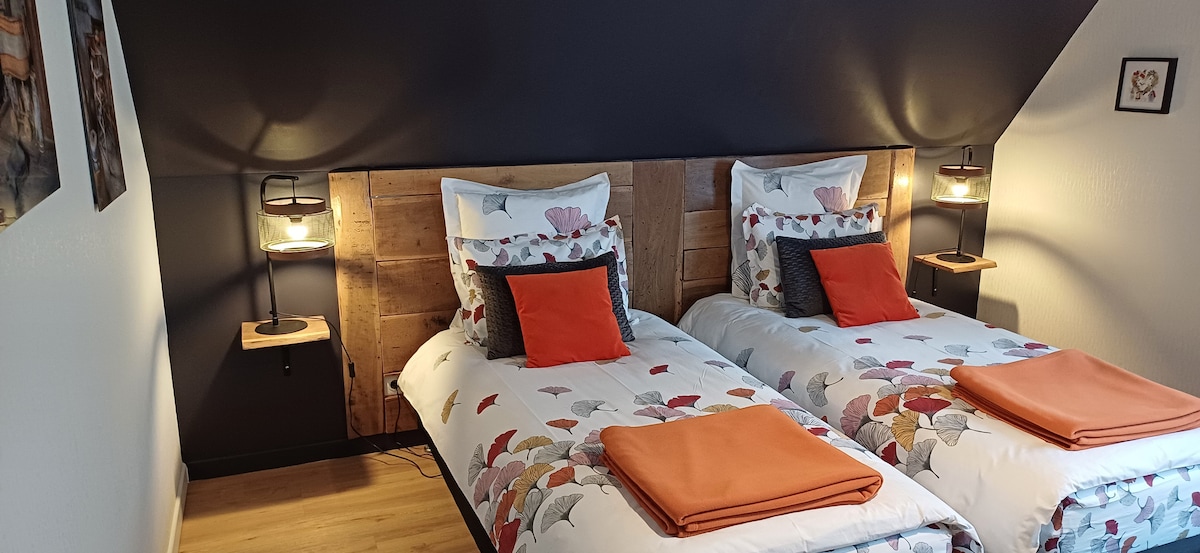 A cozy guest room is furnished with two twin beds, each dressed in floral-patterned bedding and complemented by orange accents. Wooden headboards are situated against dark-colored walls. Nightstands with lamps are positioned on either side of the beds, adding functionality and warmth to the space.