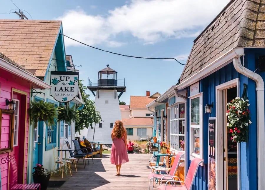 A vibrant row of cottage-style buildings in pastel colors is depicted, with a person in a pink dress walking along a wooden pathway. Decorative flowers hang from the blue and pink cottages, while a lighthouse stands in the background against a clear sky.