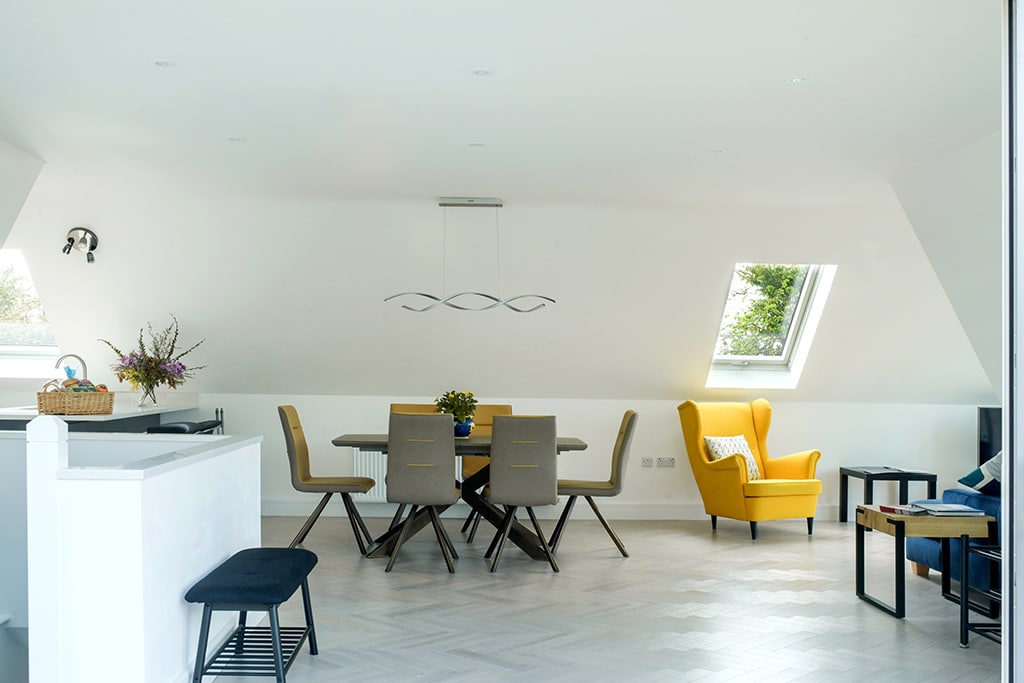 An airy open-plan living area is displayed, featuring a dining table surrounded by grey chairs. A yellow accent chair adds a pop of color, complemented by natural light from skylights above. A kitchen counter is visible on the left, with a small bench near the wall.