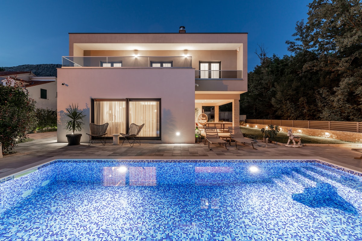 A modern villa is presented under a twilight sky, featuring a sparkling pool surrounded by outdoor seating areas. The facade includes large glass doors that lead to an inviting indoor space. Soft lighting highlights the architectural details and enhances the serene environment.
