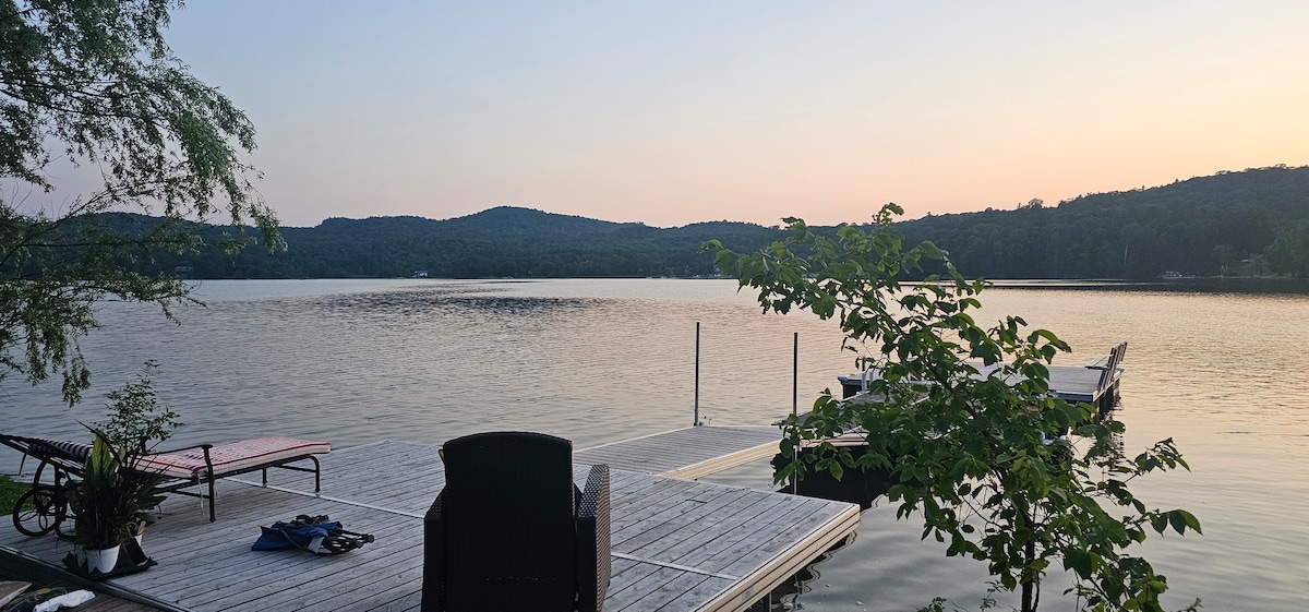 A wooden dock extends over tranquil waters, framed by gently rolling hills in the distance. Comfortable seating is positioned for relaxation, with lush greenery providing a natural backdrop. The soft hues of sunset create a serene ambiance, reflecting on the calm lake surface.