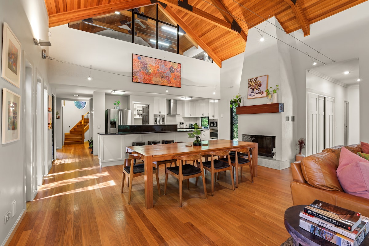 An open-plan living space features a dining table surrounded by six chairs beneath striking cathedral ceilings. Large windows allow natural light to fill the room, enhancing the view of the stylish kitchen equipped with modern appliances. A cozy seating area is visible in the foreground.