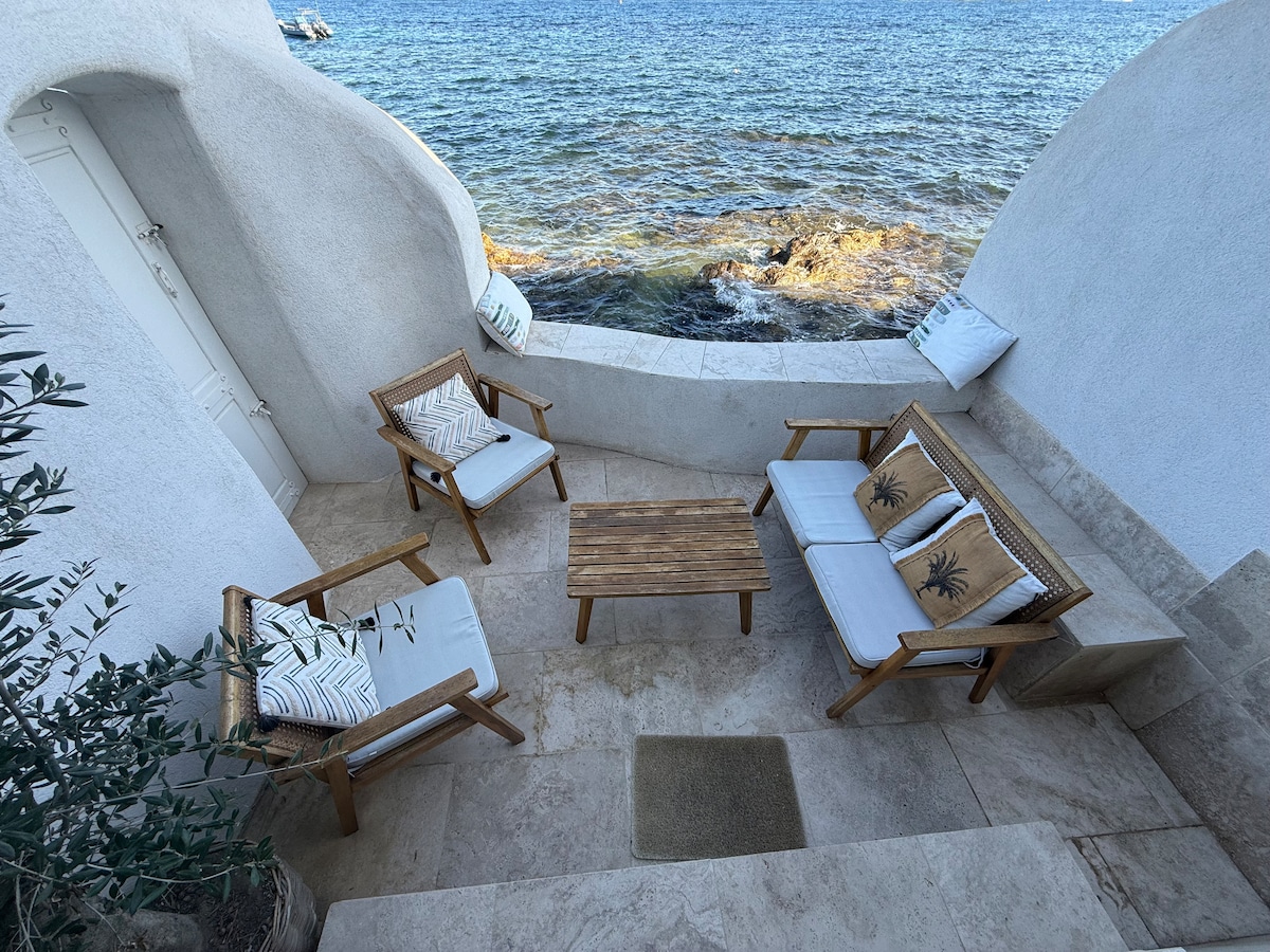An outdoor seating area is situated near the water's edge, featuring two wooden chairs and a matching table. Cushions with palm leaf designs adorn the chairs, while the surrounding walls are smoothly finished in white. The view of the sea is visible in the background.