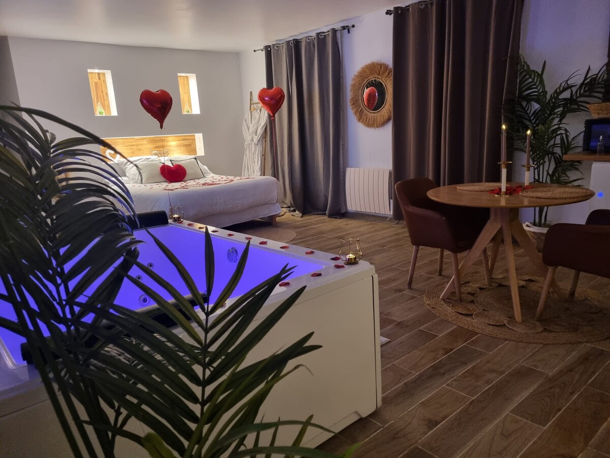 An inviting space features a cozy sleeping area with a spacious bed adorned with decorative elements. A jacuzzi with ambient lighting is nearby, complemented by a dining area with a round table and chairs. Greenery adds a touch of warmth to the setting.