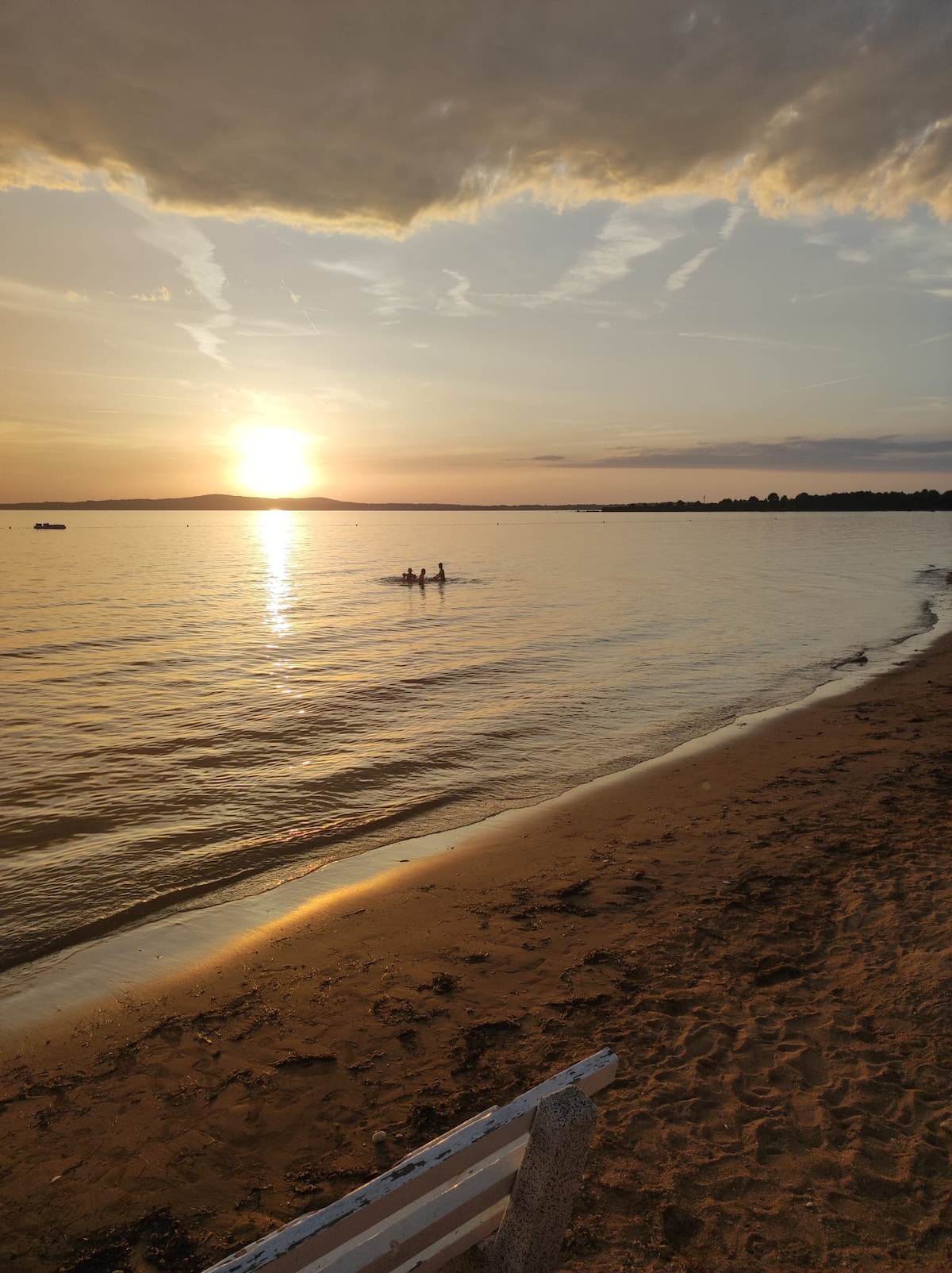 A tranquil beach scene at sunset captures soft waves lapping at the sandy shore. Silhouettes of two people are visible in the water. The sky is painted with warm hues, reflecting on the water's surface, creating a serene and calm atmosphere.