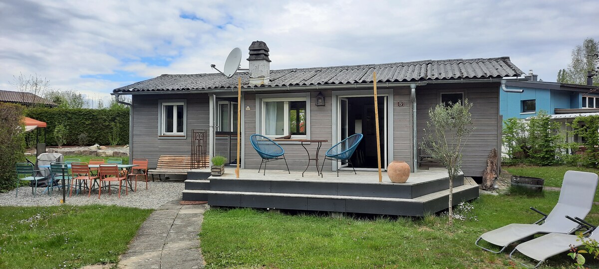A wooden chalet is featured, surrounded by greenery and a spacious lawn. A deck with two blue chairs and a small table is visible, ideal for outdoor seating. The house's exterior includes several windows, and sun loungers are placed in the grass.