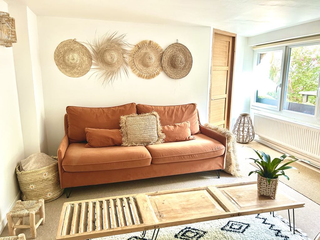 A warm living area is presented, featuring a terracotta-colored sofa adorned with textured pillows. Natural woven decor hangs on the wall, and a wooden coffee table sits in front, complemented by a potted plant. A large window invites ample daylight, highlighting the serene ambiance.