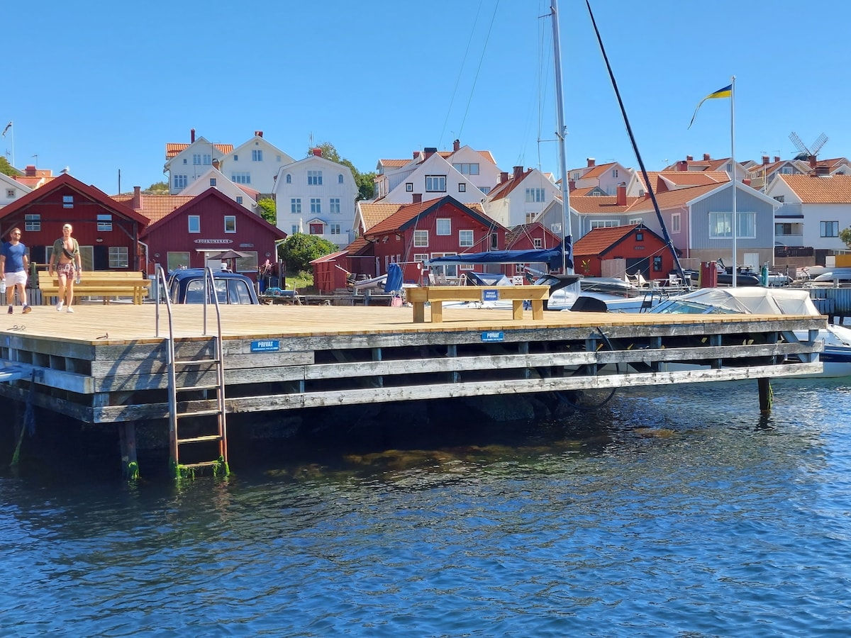 A wooden pier extends over calm waters, featuring a single staircase leading down to the water. Several benches provide seating for visitors. Colorful buildings are visible in the background, along with boats moored at the edge, creating a serene coastal atmosphere.