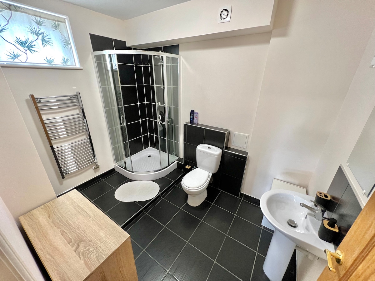 A modern bathroom features a circular glass shower enclosure beside a toilet and a sink. A heated towel rail is mounted on the wall. The floor is tiled in dark hues, and natural light enters through a small window, enhancing the clean and functional design.