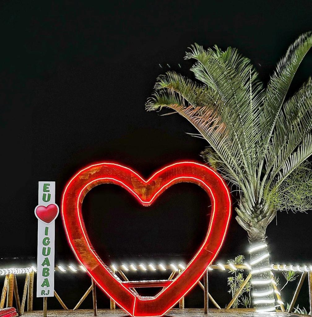 A large illuminated heart sculpture stands prominently against a night sky, surrounded by palm trees. The structure's vibrant red glow contrasts with the dark backdrop, while white lights are visible along the pathway, enhancing the inviting atmosphere of the location.