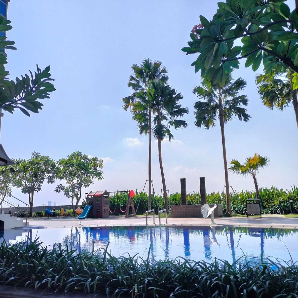 A tranquil pool area is surrounded by lush greenery and palm trees. A children's playground with colorful equipment is visible nearby, enhancing the inviting outdoor space for families. Clear blue skies reflect off the surface of the pool, contributing to a serene atmosphere.