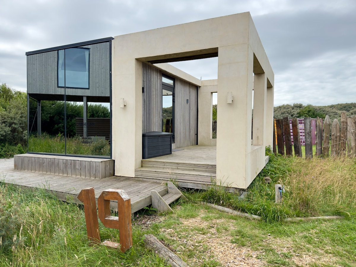 A modern eco-friendly tiny house features a combination of sleek concrete and wooden elements, surrounded by greenery. Large windows invite natural light into the interior spaces, while a wooden deck provides an outdoor area. Rustic wooden fencing and paths complement the natural landscape.