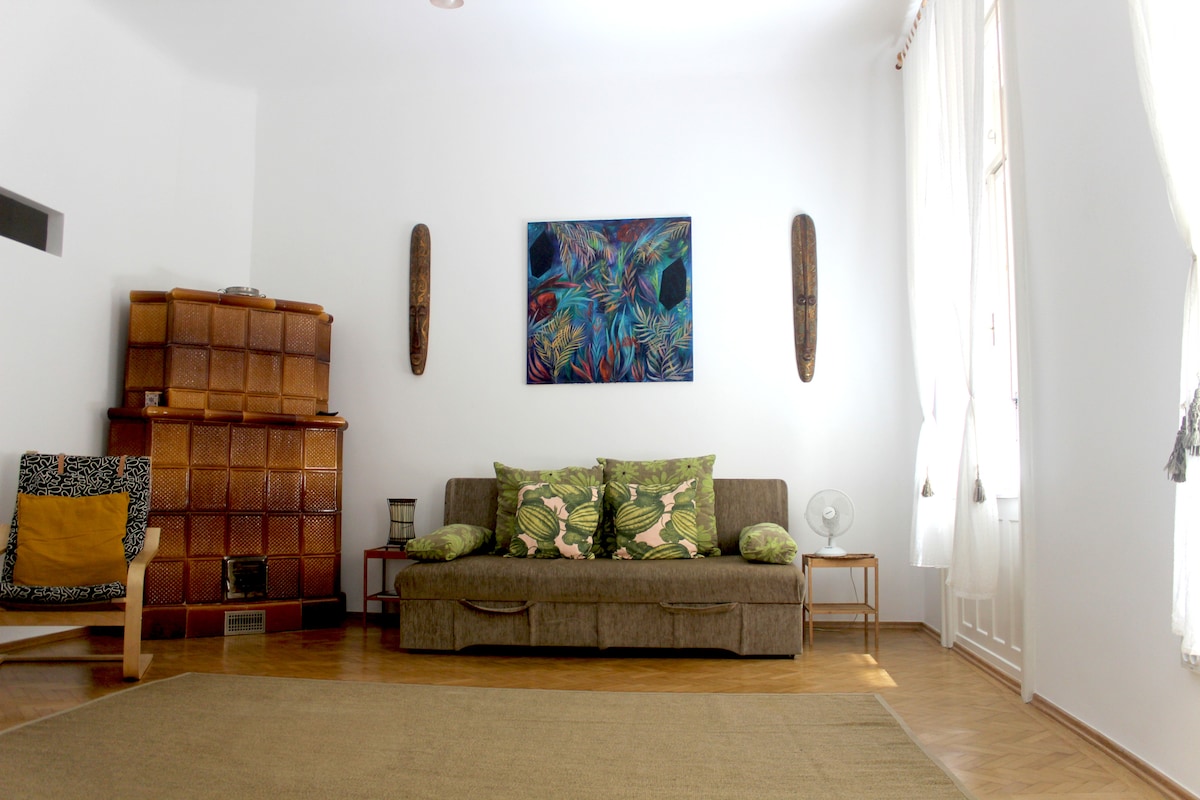 A spacious living room features a comfortable sofa adorned with green and patterned cushions. Light-colored walls are complemented by wooden flooring and a large area rug. A decorative tile stove is positioned against one wall, while a colorful piece of wall art adds interest. Windows are dressed with sheer white curtains.