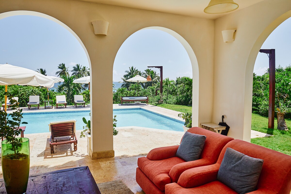 A spacious lounge area sits beneath arched openings, with lush greenery visible beyond. A turquoise pool glimmers under the sun, surrounded by sun loungers and umbrellas. Cozy orange sofas offer a relaxed seating option, enhancing the inviting ambiance of the outdoor retreat.