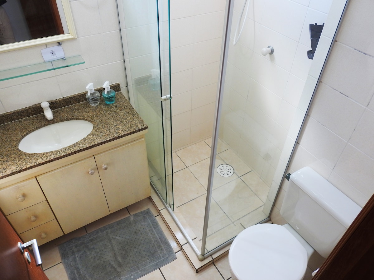 A well-organized bathroom is displayed, featuring a shower stall with clear glass doors. A modern sink is positioned on a granite countertop with storage drawers below. A toilet is placed adjacent, and a mat lies on the tiled floor, emphasizing the clean and functional space.