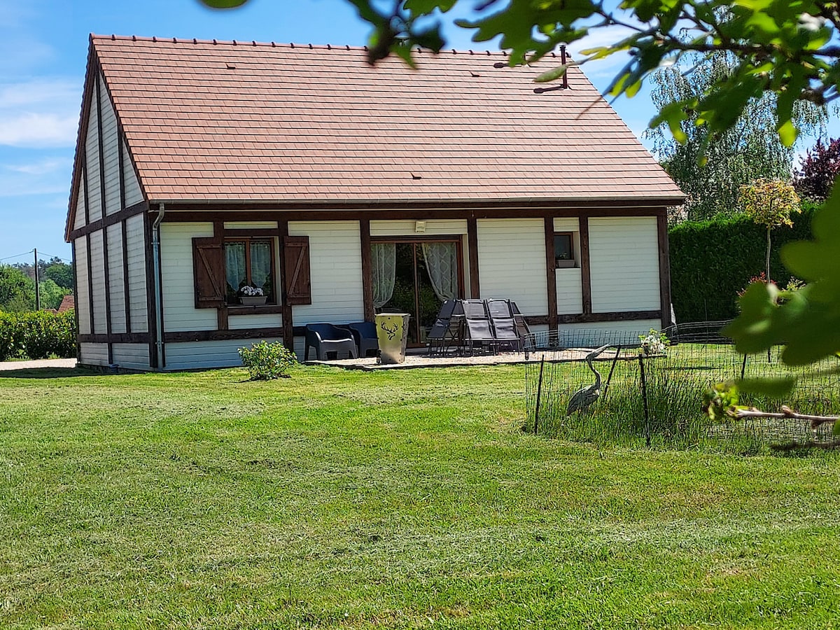 A single-story house is situated in a lush green landscape, featuring a spacious terrace adorned with outdoor seating. Neutral exterior colors complement the tiled roof, while large windows allow natural light into the living space. Surrounding greenery provides a serene atmosphere.