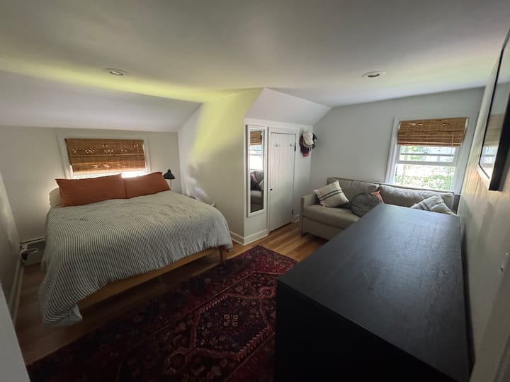 Upstairs bedroom with a queen bed and couch. Dresser and small closet. TV mounted on the wall. Window unit AC in summer