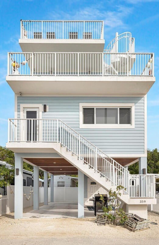 A multi-level vacation rental is shown, featuring a light blue exterior and a rooftop deck enclosed by white railings. A staircase provides access to the raised entrance, with a covered area beneath for additional shade. The surroundings are well-maintained, contributing to a relaxing environment.