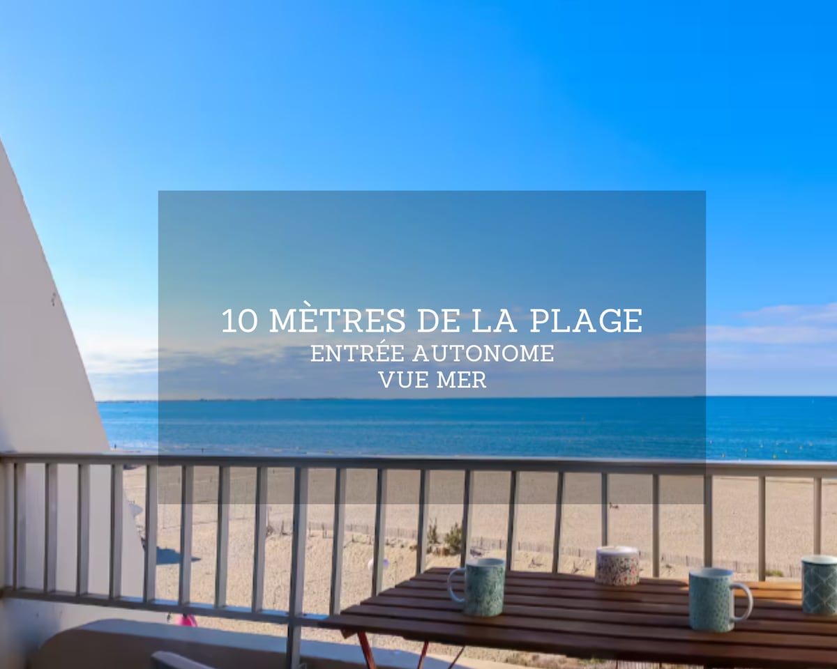 A balcony view presents the beach only 10 meters away, with expansive blue skies above and calm sea waters stretching to the horizon. A wooden table is seen, accompanied by several cups, creating a peaceful outdoor space for relaxation.