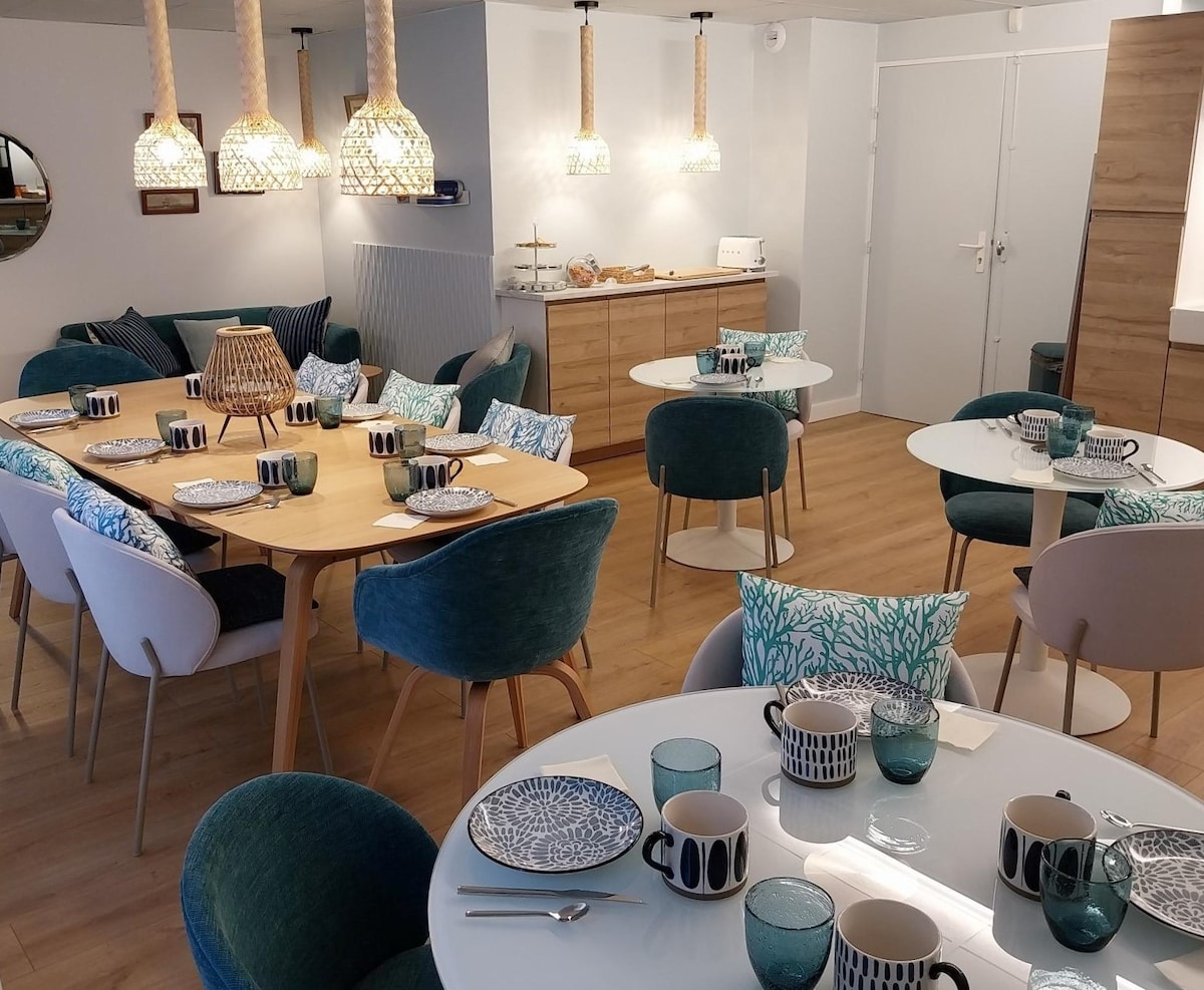 A dining area is arranged with multiple tables, showcasing a mix of circular and rectangular surfaces. Comfortable chairs in various shades surround the tables, adorned with decorative pillows. Elegant pendant lights hang overhead, creating a welcoming space for shared meals.