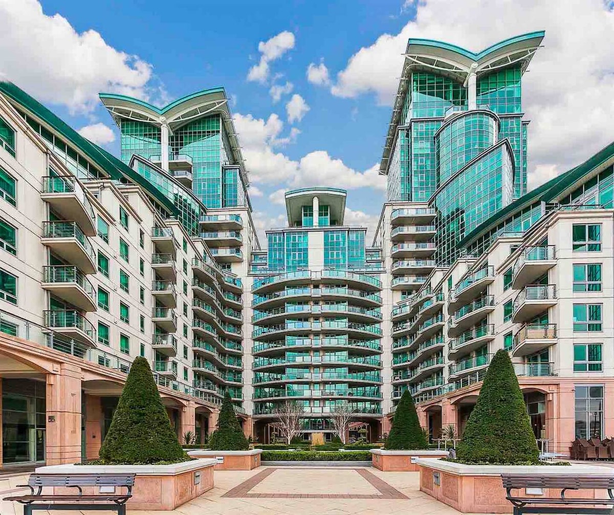 An expansive view of a modern residential building complex is presented, showcasing sleek, glass-fronted structures with distinctive architectural features. The courtyard below is landscaped with manicured hedges and pathways, framed by greenery and sunlight filtering through scattered clouds.