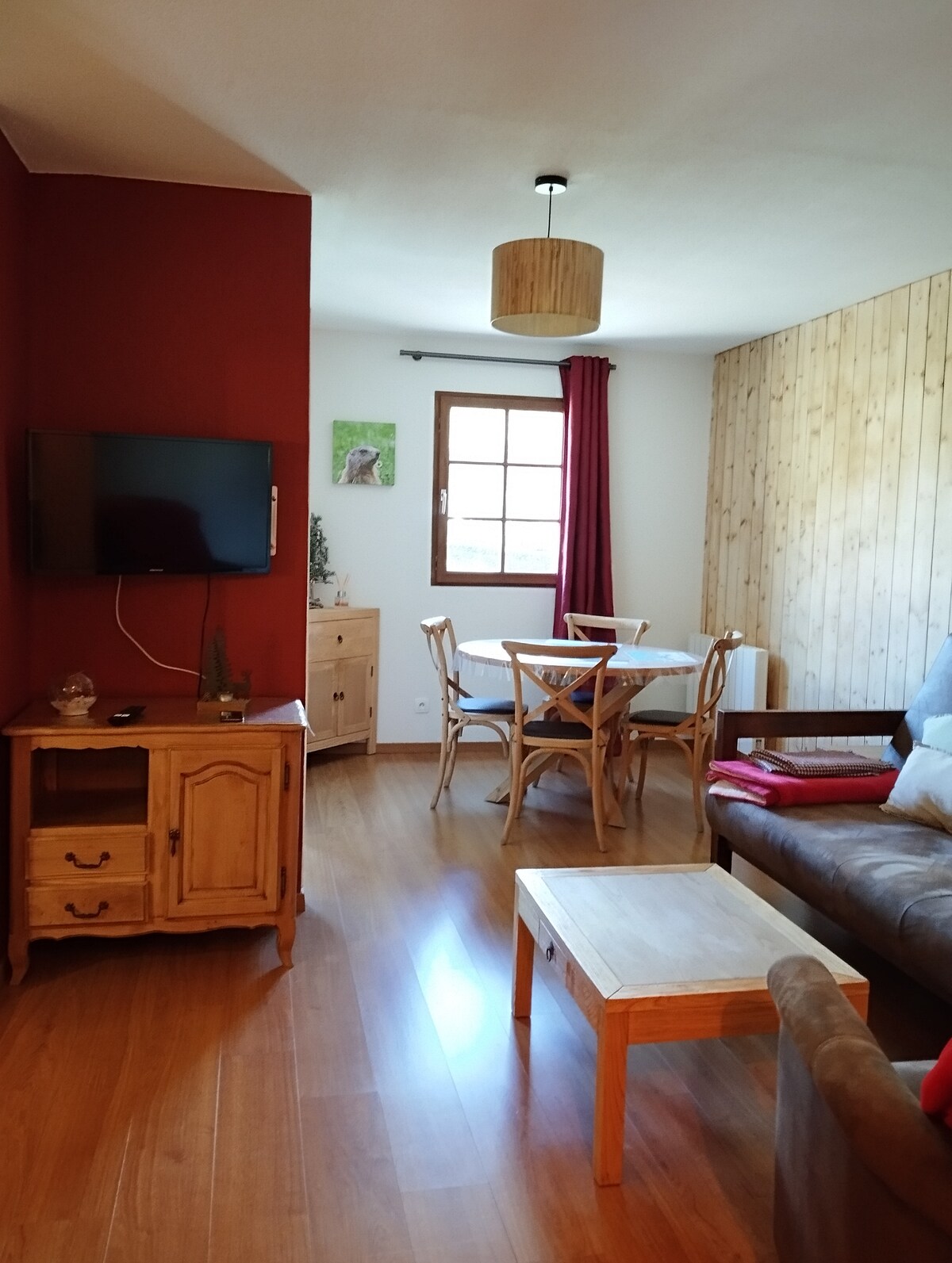 A bright living area features a wooden coffee table and a cozy sofa, complemented by a wooden entertainment unit with a television. A dining table with four chairs sits nearby, while large windows provide natural light. The walls display a mix of red paint and wooden panels.