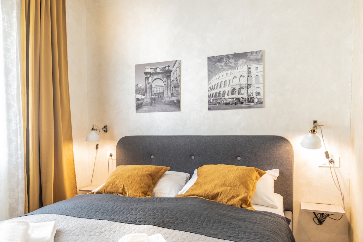 A cozy sleeping area features a comfortable bed with gray bedding and gold accent pillows. Two black-and-white framed photographs adorn the walls, depicting architectural landmarks. Soft, natural light is filtered through sheer curtains, and bedside lamps provide illumination.