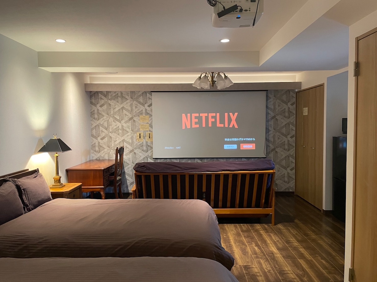 This room features two semi-double beds and a sofa bed, providing versatile sleeping arrangements. A large projector screen displays the Netflix logo, while a wooden desk and lamp add functional workspace. Natural light may enter through the partially covered window, enhancing the relaxing environment.