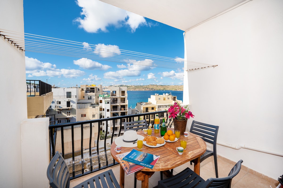 A terrace features a wooden table surrounded by black chairs, adorned with a colorful flower arrangement and fresh fruit. The background showcases views of the sea and nearby buildings under a clear blue sky dotted with clouds.