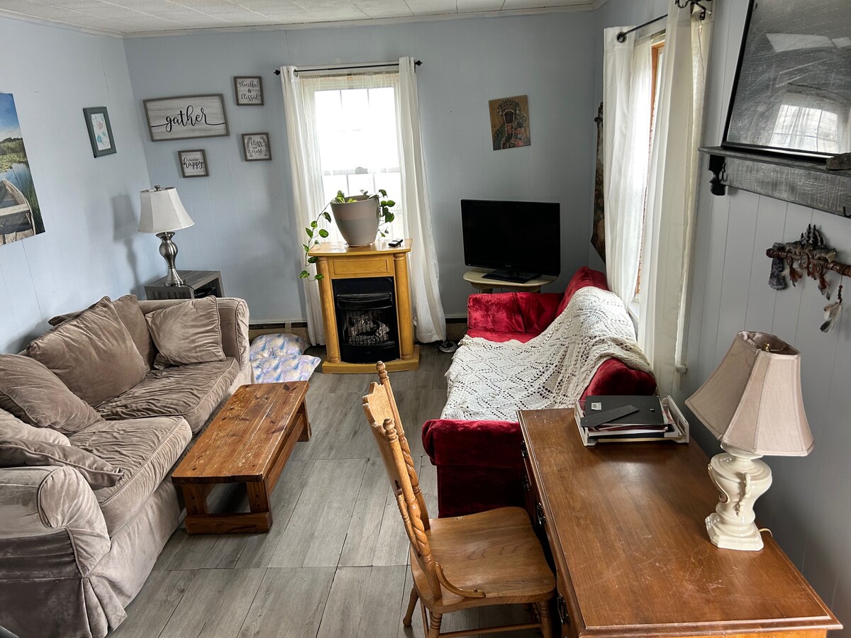 The cozy living area features a soft sofa and loveseat, creating a relaxed space. A wooden coffee table is placed centrally, while a small desk with a lamp occupies one corner. A television is positioned near a faux fireplace, with light streaming through a nearby window.