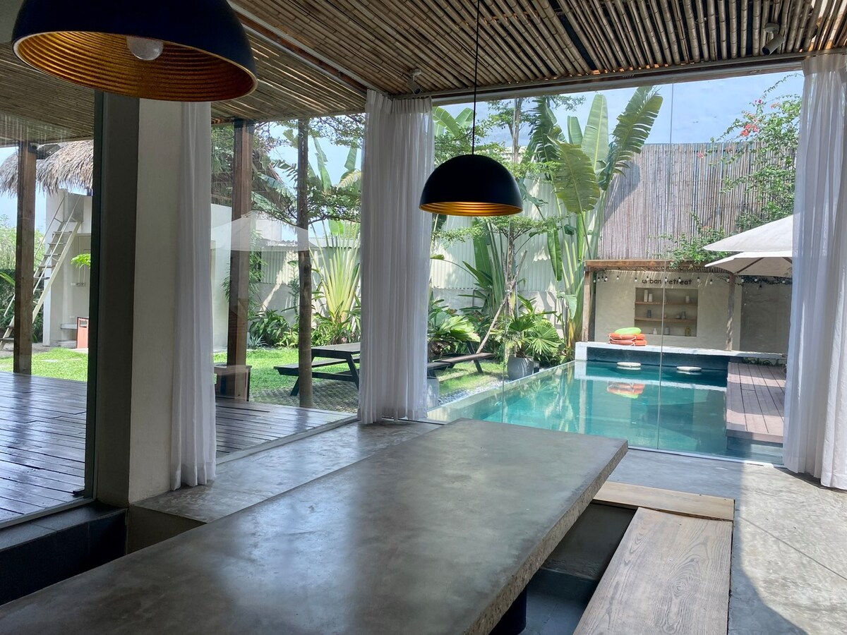 An open dining space features a concrete table, complemented by pendant lighting. Large glass windows offer views of a lush garden and an adjacent swimming pool, enhancing the connection with nature. Natural light floods the area, creating a bright and refreshing ambiance.
