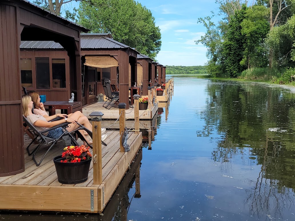 Two individuals relax in zero-gravity chairs on a wooden deck, surrounded by serene waters, with cabins visible in the background. The deck is adorned with blooming flowers in planters, and the lush greenery and trees line the opposite shore, enhancing the natural setting.