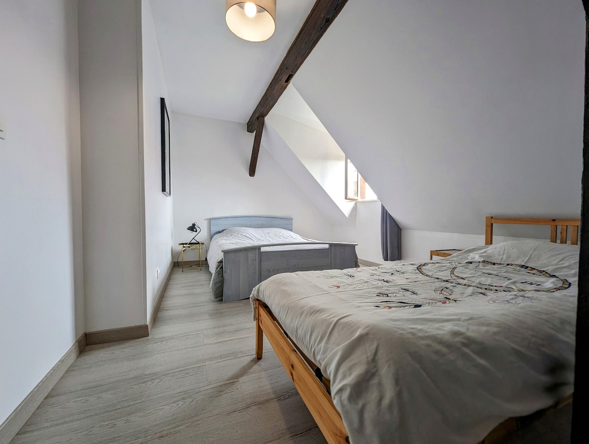 The sleeping area features two beds positioned under sloped ceilings, with a spacious wooden bed frame alongside a smaller bed. Natural light filters through a window, illuminating the light-colored walls and wooden accents, creating a serene and bright environment.