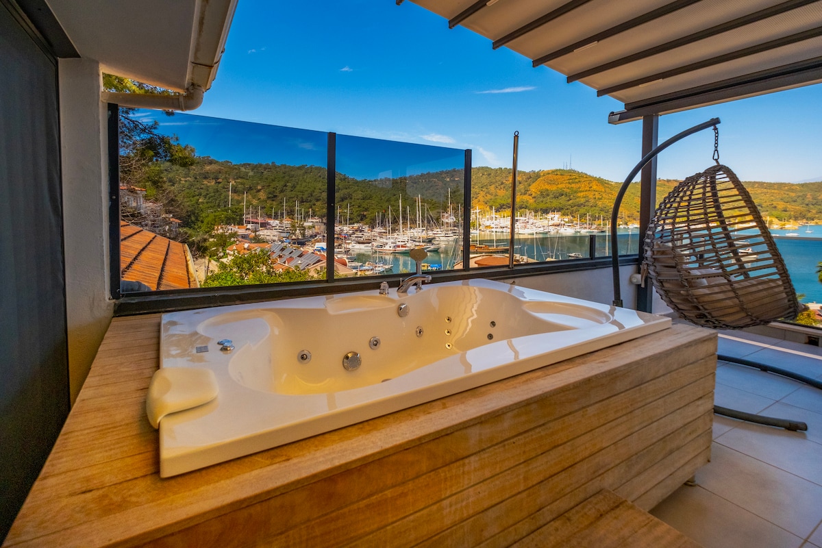 A spacious hot tub is situated on a wooden deck, offering views of the marina and hills in the background. A hanging chair is positioned nearby, providing a relaxed seating option. Large glass panels enhance the panoramic perspective of the sea and surrounding nature.