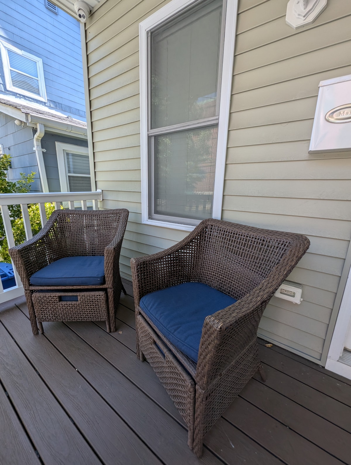 A front porch is furnished with two woven chairs featuring blue cushions. The wooden flooring complements the light-colored siding of the house, while a window allows natural light to brighten the space.