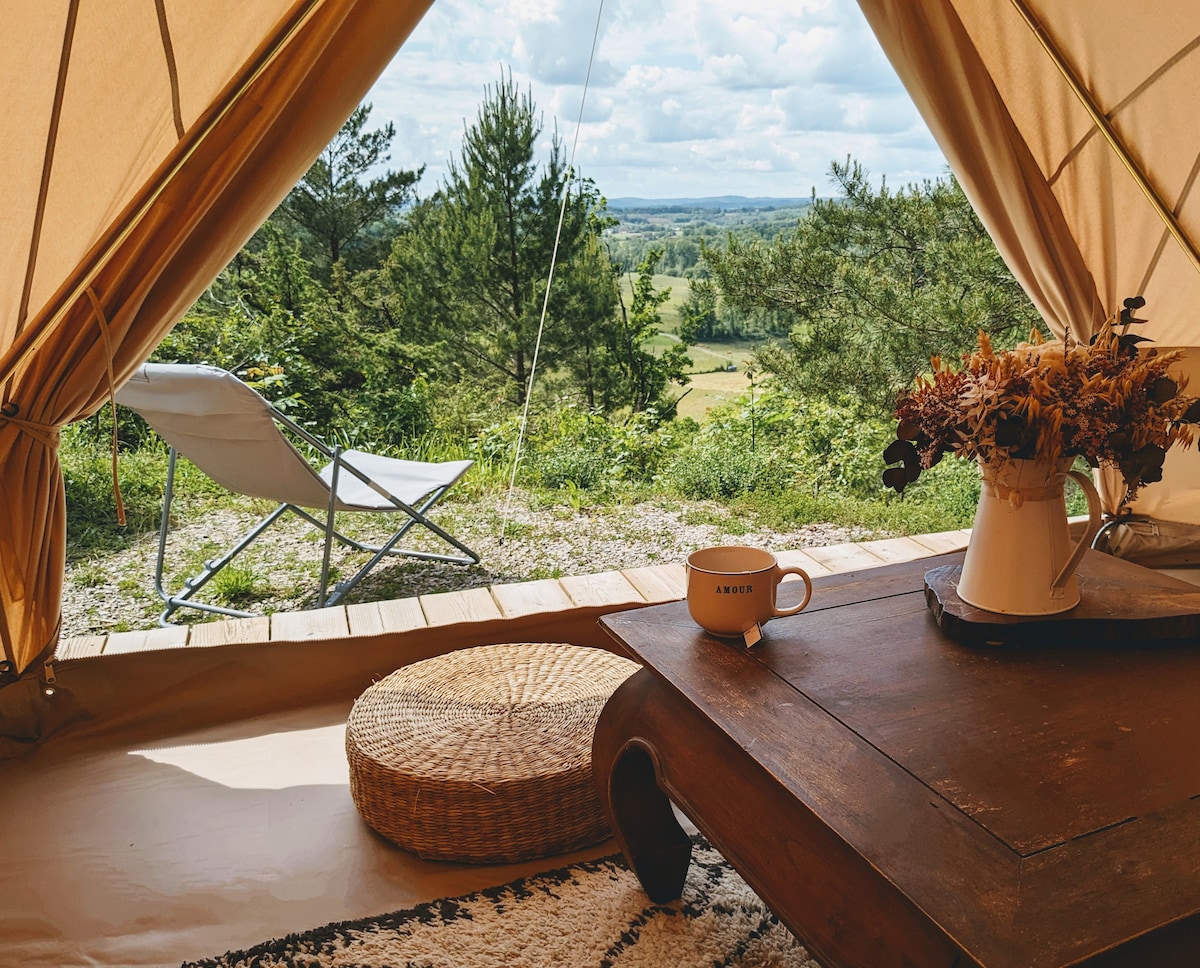 The interior of the tipi showcases a cozy seating area with a low wooden table and a woven pouf. A view of the lush countryside is seen through the entrance, with a folding chair positioned outside, inviting relaxation surrounded by nature.