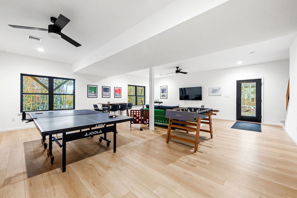 The game room features a ping-pong table, foosball table, and a cozy seating area. Large windows allow for ample natural light, and the wooden flooring adds a warm touch. A flat-screen TV is mounted on the wall, enhancing the entertainment options.