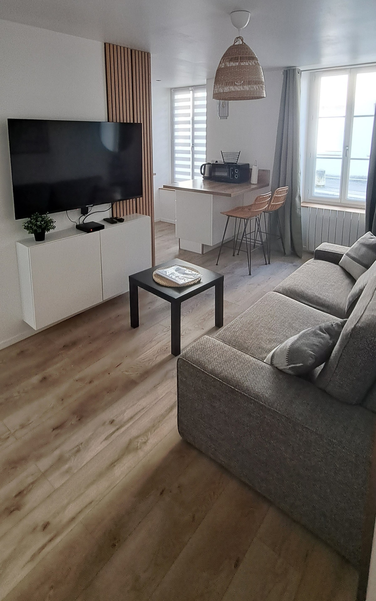 A living area features a grey sofa and a coffee table, with a wall-mounted television positioned across from the seating. A dining space with three stools is visible, alongside large windows that allow natural light to fill the room.