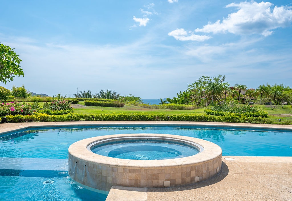 A circular hot tub is positioned within a large pool, surrounded by a stone deck. Lush green landscaping extends towards the ocean in the background, under a clear blue sky with some clouds.