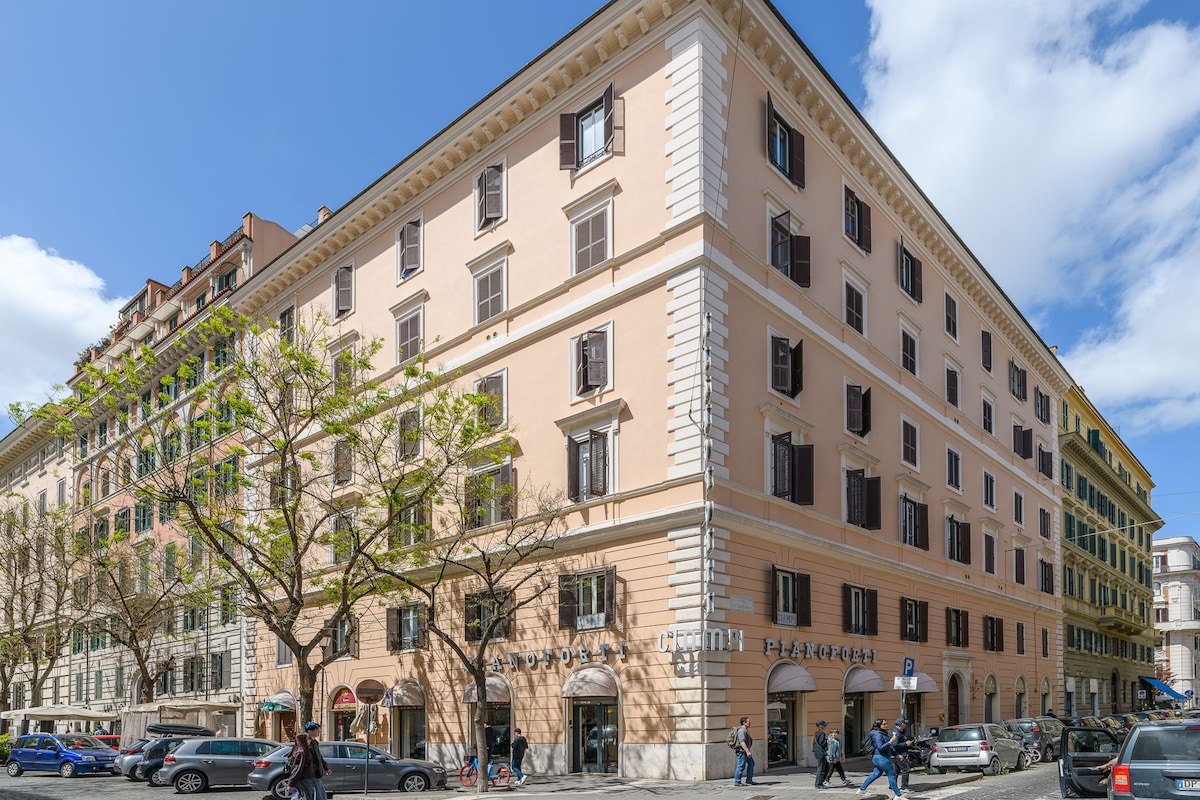 A modern building with classic architectural details is viewed from the street. Large windows are symmetrically arranged across the facade, and trees line the sidewalk, providing a glimpse of daily life in the bustling city. Clouds drift across a blue sky overhead.