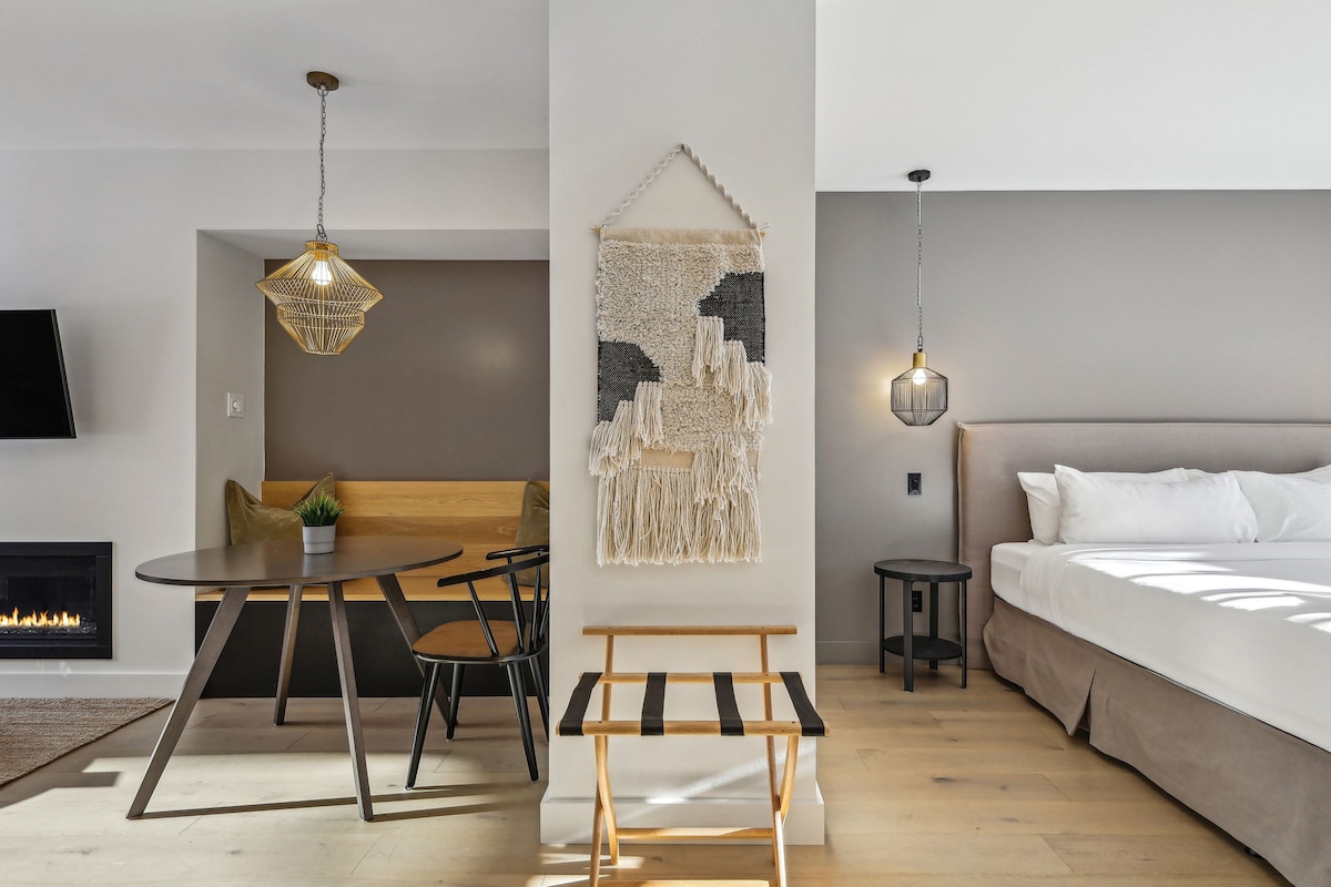 A modern interior features a spacious bedroom area with a soft bed dressed in white linens. A stylish table and chairs are positioned nearby, complemented by a decorative wall hanging. A cozy nook accentuates the space with a built-in bench and greenery.