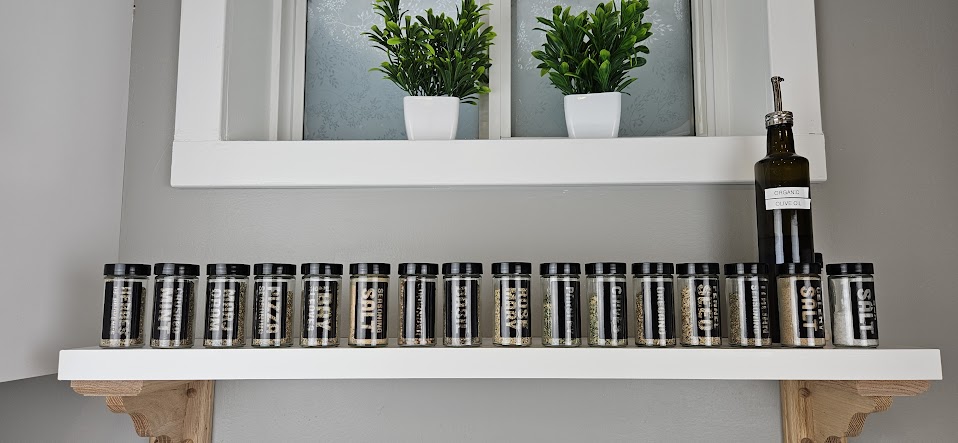A clean, modern shelf displays an array of jars labeled with various spices. Small potted plants add a touch of greenery above the jars, while a decorative oil dispenser complements the rustic appeal. The background features a softly textured wall in light gray.
