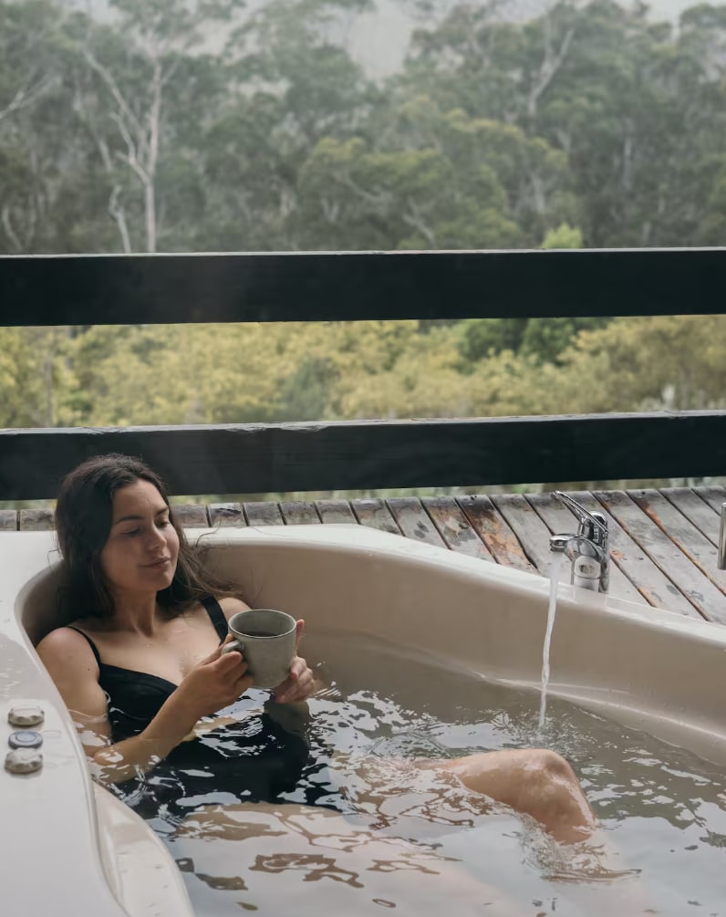 An outdoor spa tub is positioned on a wooden deck, overlooking lush greenery. Water flows gently from a spout into the tub, creating a serene atmosphere. A person is relaxing in the tub while holding a cup, showcasing the tranquil experience available.