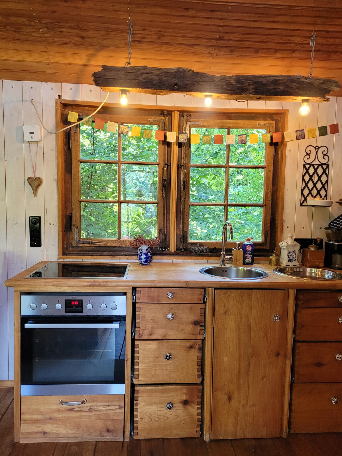 A rustic kitchen area features wooden cabinetry with a built-in oven and a stainless-steel sink. Natural light fills the space through two windows adorned with decorative garlands, revealing lush greenery outside and enhancing the cozy ambiance.