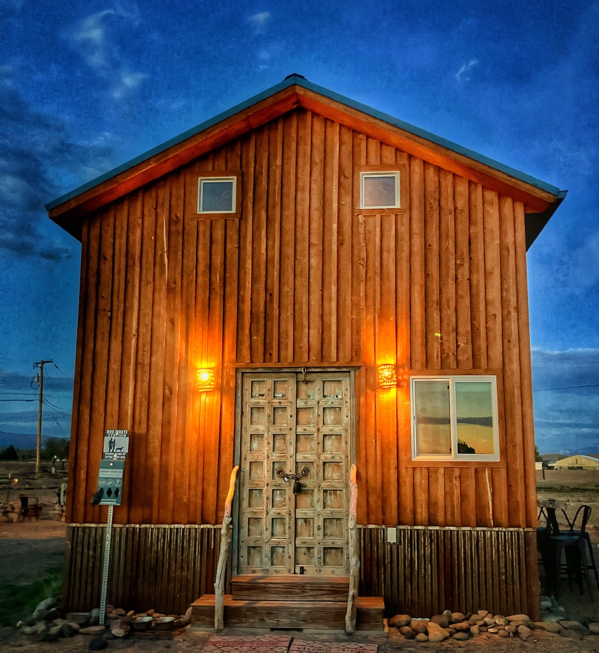 The exterior of the tiny home showcases a rustic wooden facade, complemented by warm lighting on either side of the entrance. A large, decorative door with intricate designs invites guests inside, while a single window provides a glimpse of the interior.