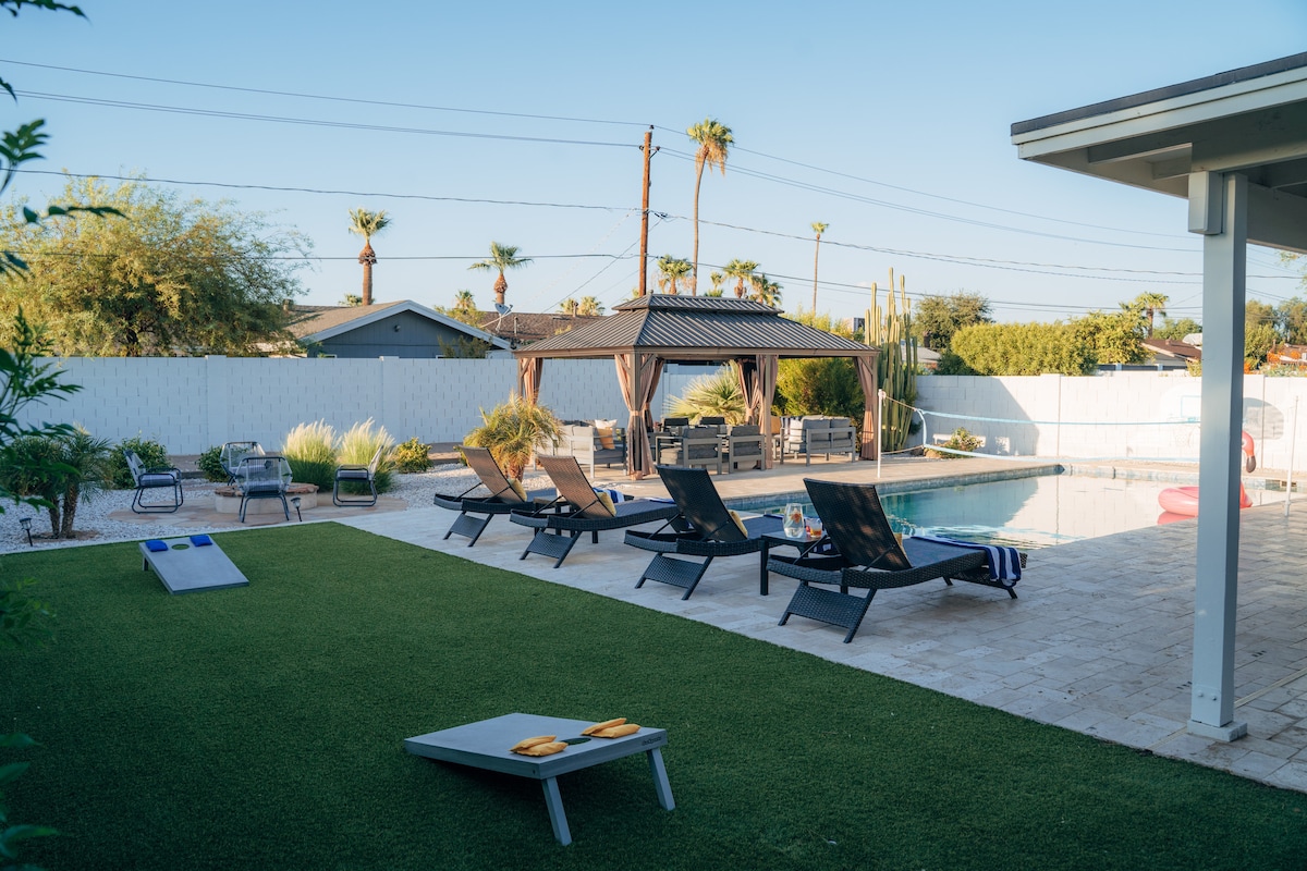 A spacious outdoor area features a heated pool surrounded by lounge chairs and a gazebo. Near the pool, game equipment such as a cornhole set is visible on the artificial grass. Lush landscaping and tall palm trees add to the inviting environment.