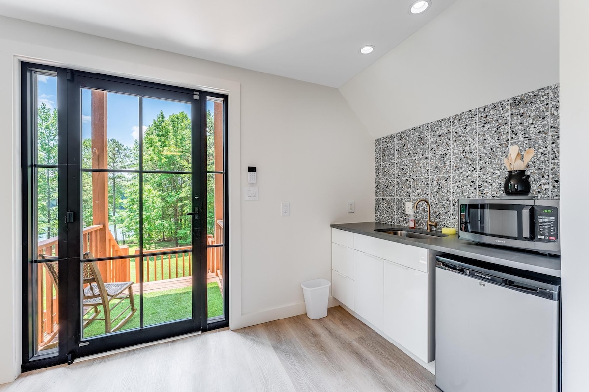 A bright kitchenette is seen with modern stainless-steel appliances, including a microwave and refrigerator. The textured backsplash offers a unique design element. Large glass doors open to a scenic deck overlooking greenery, enhancing the connection to the outdoor landscape.