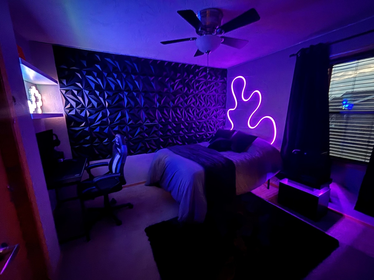 A modern bedroom design features a textured black accent wall complemented by a vibrant neon light. A spacious bed is placed against the wall, while a comfortable chair and desk are situated nearby. Natural light filters through a window covered by dark curtains.
